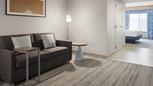 A modern hotel room with a grey sofa, a small side table, a floor lamp, and a bed with a view of the city. - Hilton Garden Inn Albuquerque Airport (Albuquerque)