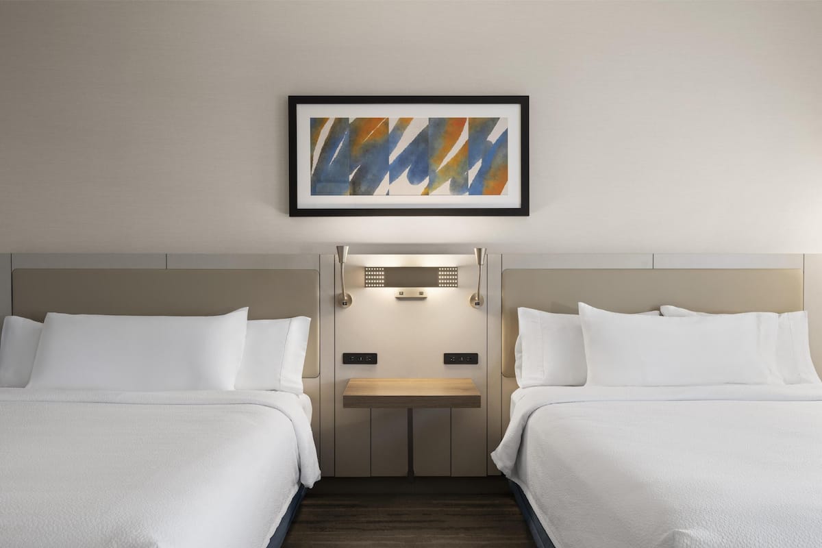 A hotel room with two beds, a wooden nightstand, a wall-mounted light, and a framed abstract painting.