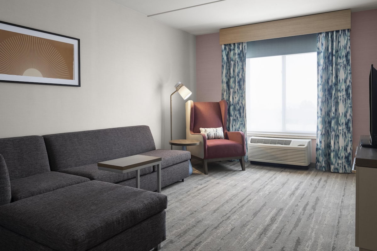 A modern hotel room with a grey sofa, a red armchair, a small table, a floor lamp, and a large window with curtains.