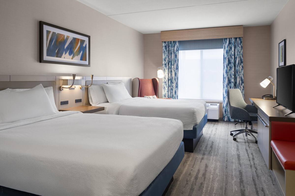 A hotel room with two beds, a desk with a chair, a TV, and a window with curtains.