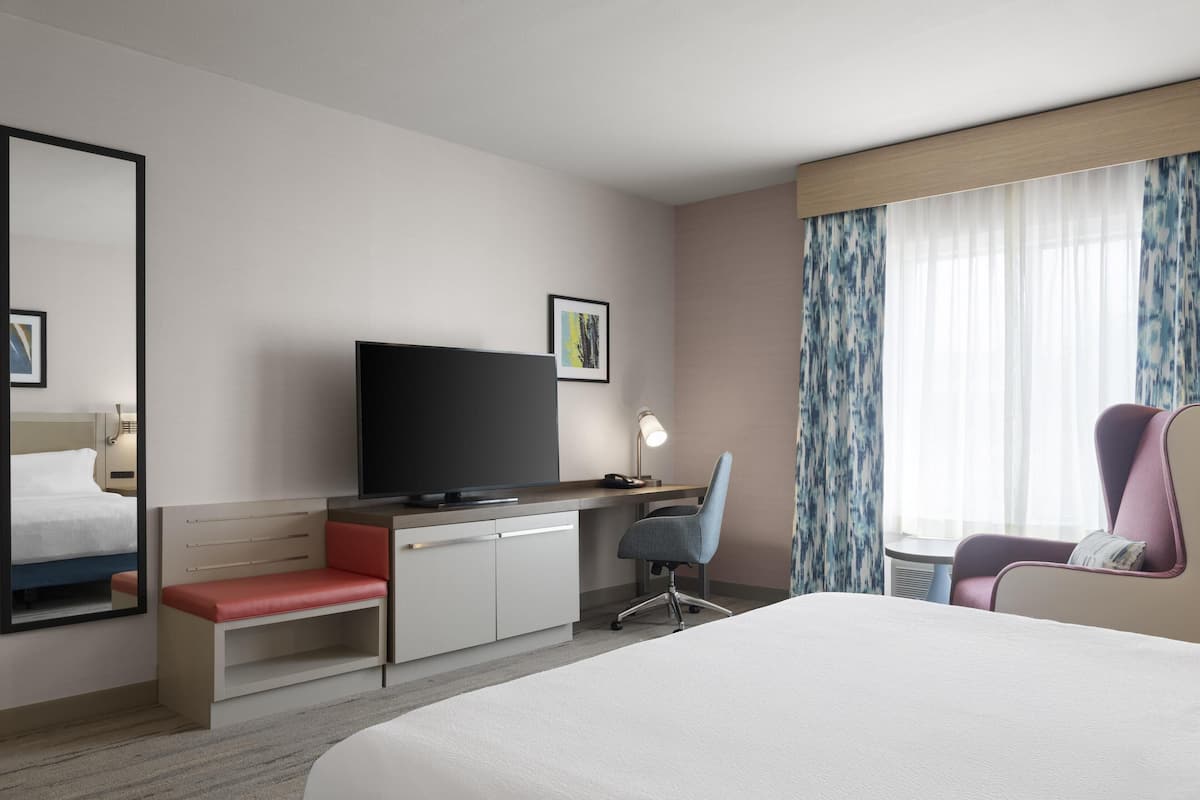 A hotel room with a bed, a desk with a computer, a chair, a TV, and a window with curtains.