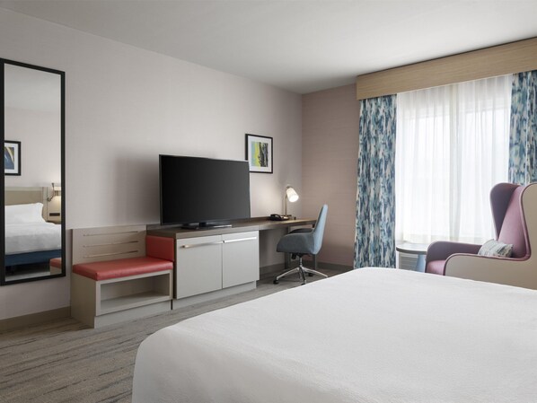 Une chambre d’hôtel avec un lit, un bureau avec un ordinateur, une chaise, une télévision et une fenêtre avec des rideaux.