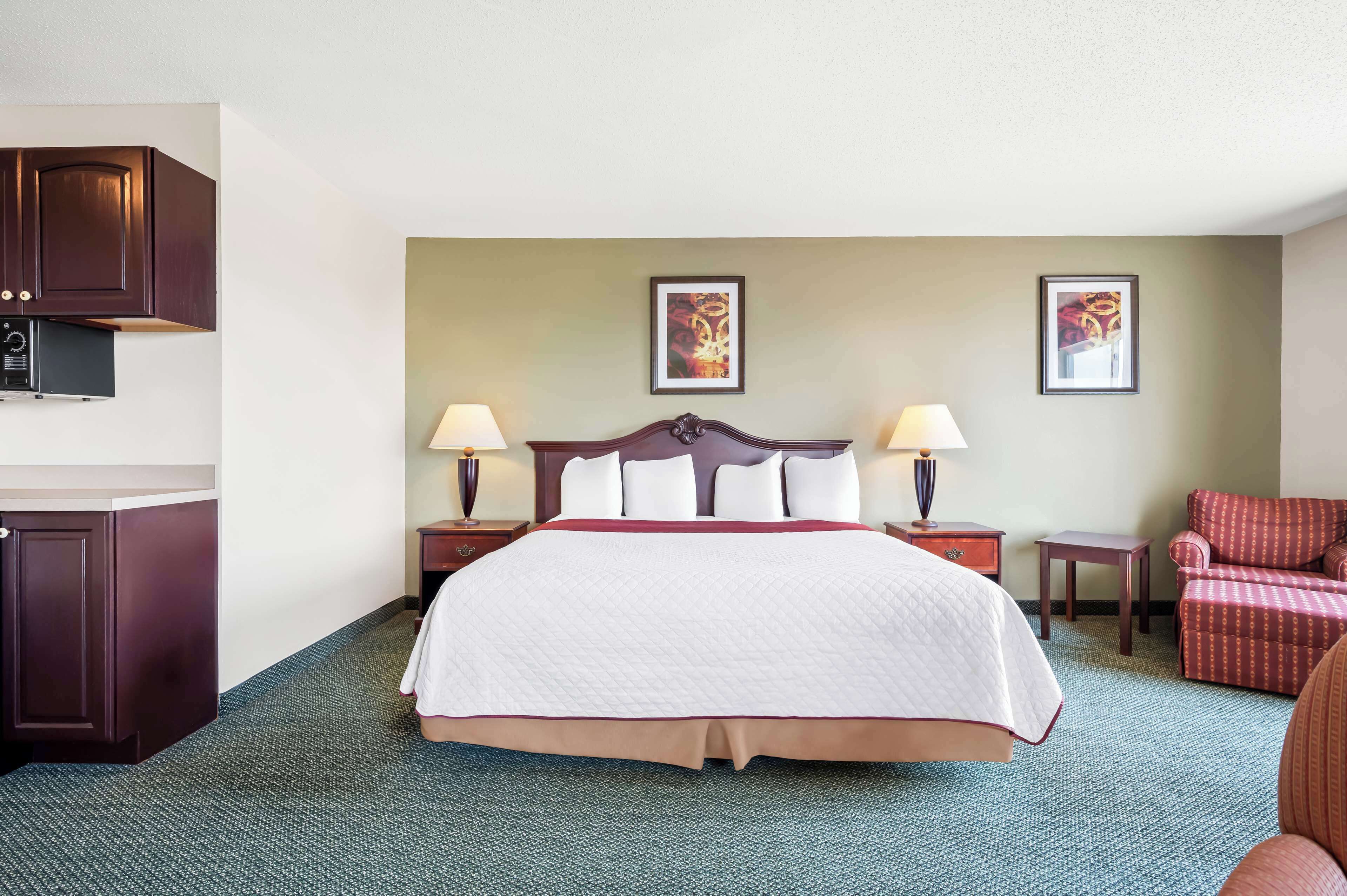 Una habitación de hotel con una cama grande, dos mesitas de noche con lámparas, un sillón rojo y una kitchenette con microondas.