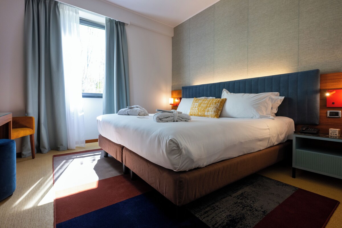 Une chambre d’hôtel avec un grand lit, une table de chevet, un bureau et une fenêtre avec des rideaux.