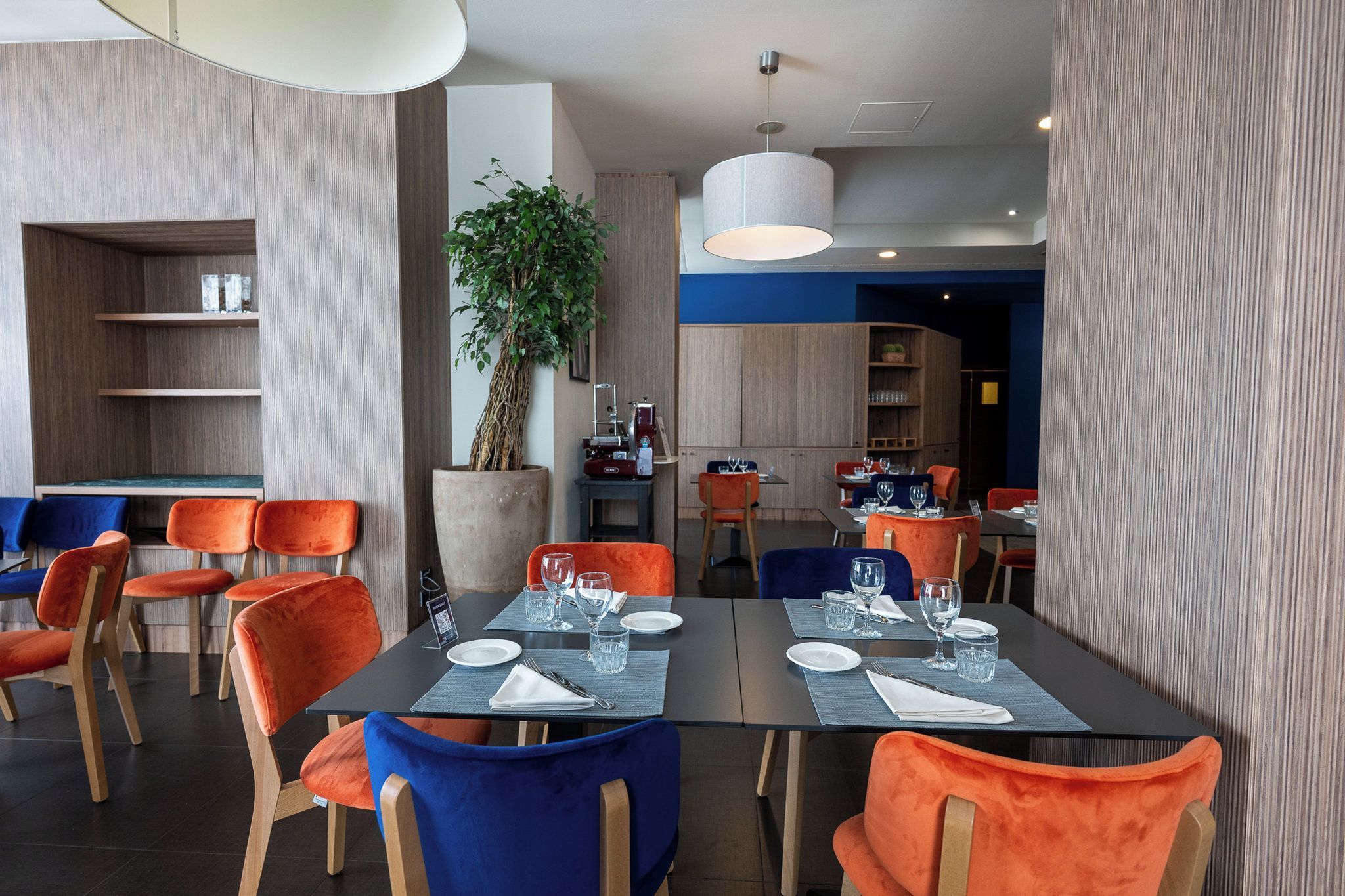 A modern dining area with tables set for two, featuring a mix of orange and blue chairs.