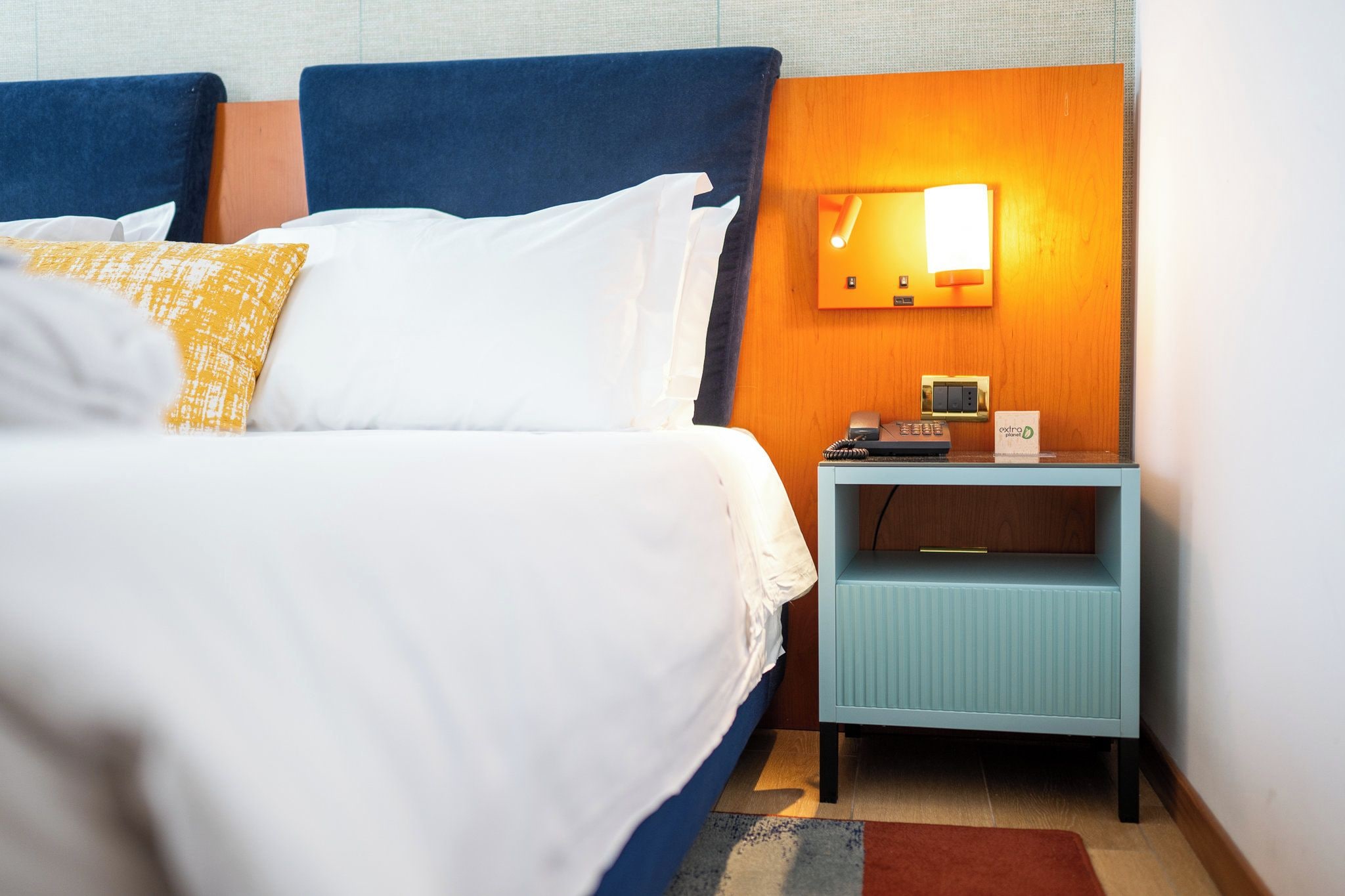 A hotel room with a bed, a nightstand with a telephone, and a wall-mounted light.