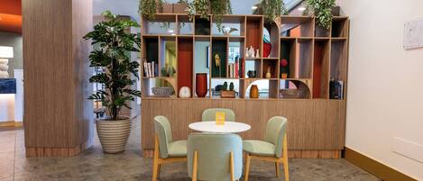 Un área de recepción con un mostrador de madera, dos sillas, una planta grande en maceta y una estantería con varios objetos decorativos.