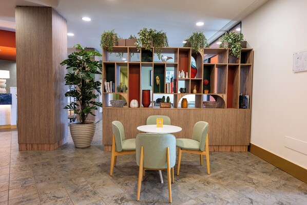 Un espace d’accueil comprenant un comptoir en bois, deux chaises, une grande plante en pot et une bibliothèque contenant divers objets décoratifs.