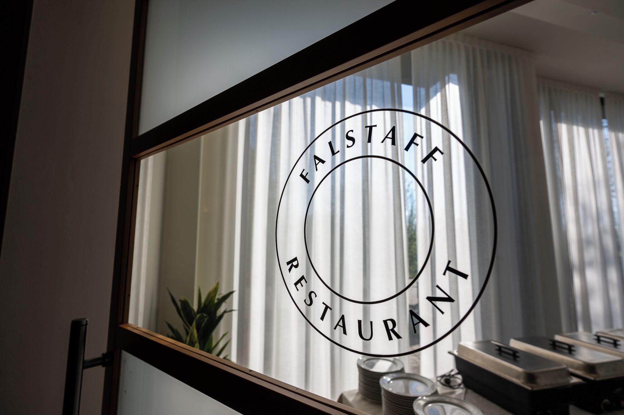 Un mirall amb un logotip circular que diu ‘FALSTAFF RESTAURANT’ i el reflex d’una habitació amb una planta i una taula amb plats.
