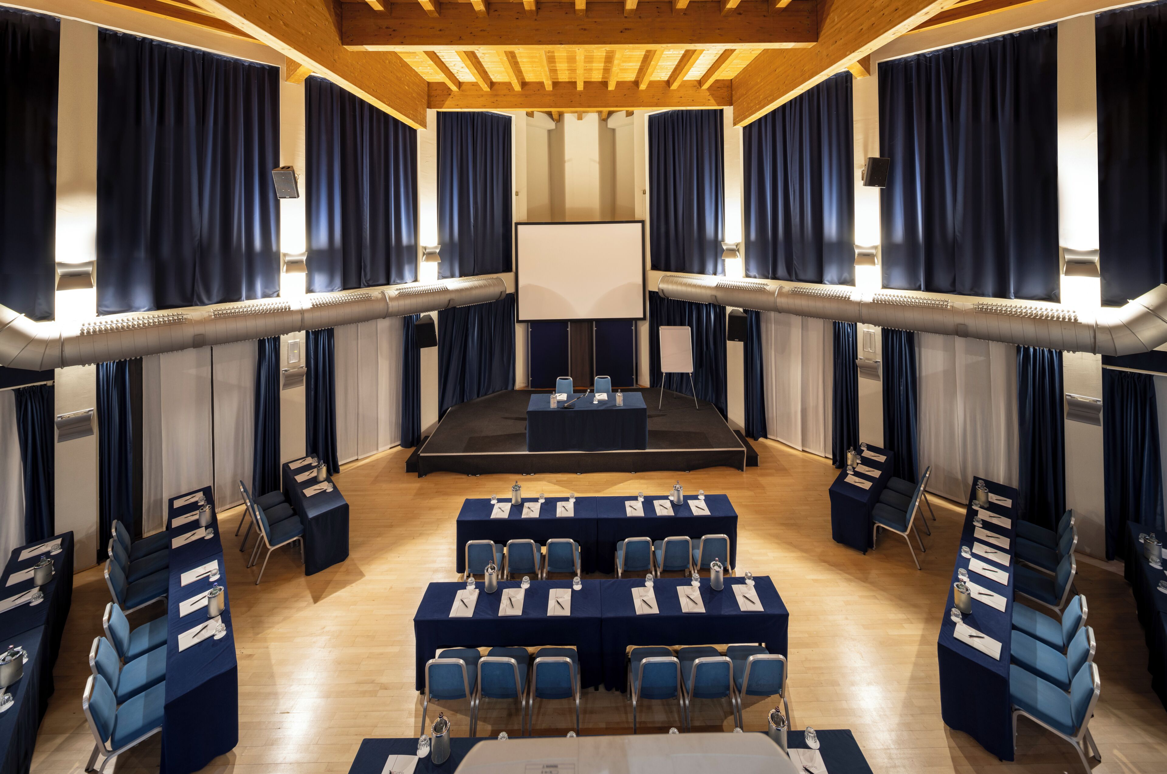 Una sala conferenze con un palco, file di tavoli e sedie, e un grande schermo.