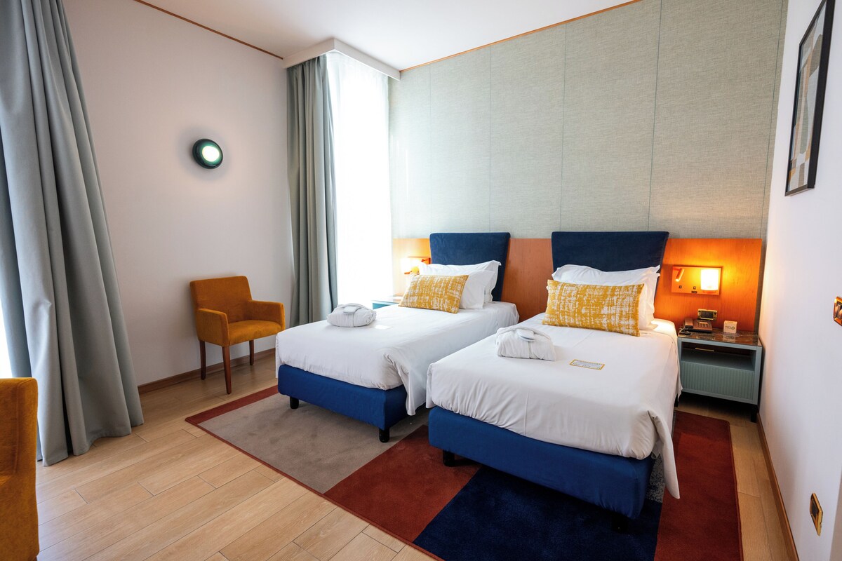 Une chambre d’hôtel avec deux lits, une chaise, une table de chevet et une fenêtre avec des rideaux.