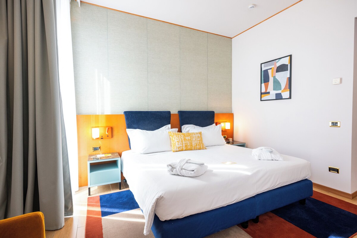 Une chambre d’hôtel avec un grand lit, deux tables de chevet, une petite table, une chaise bleue, un tapis rouge et bleu, et un tableau accroché au mur.