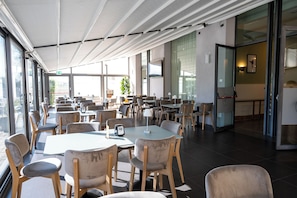 A spacious indoor dining area with multiple tables and chairs, large windows, and a modern ceiling design. - voco Parma Congressi by IHG (Parma)
