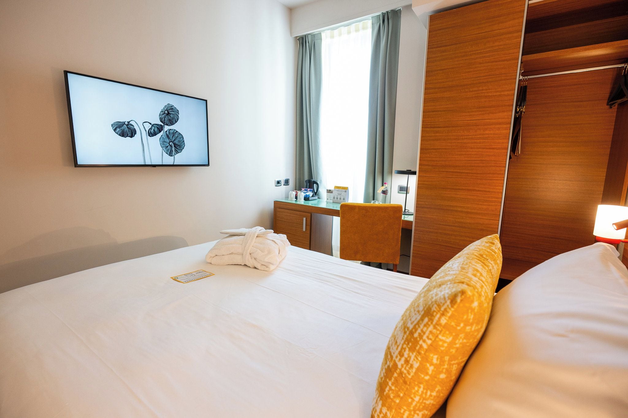 A hotel room with a bed, a desk, a chair, a wardrobe, a framed picture on the wall, and a window with curtains.