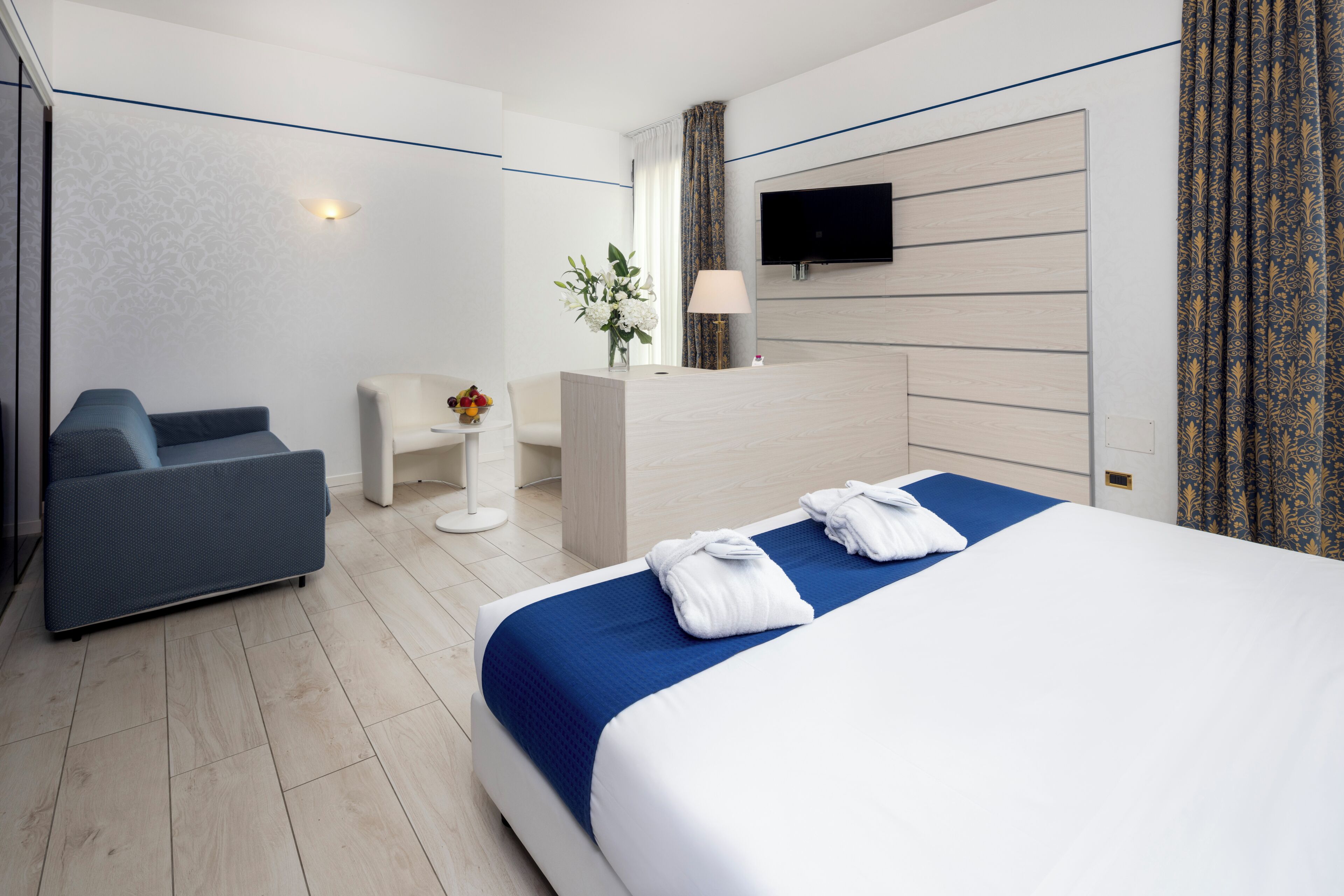 A hotel room with a bed, a sofa, a desk, a TV, and a vase with flowers.