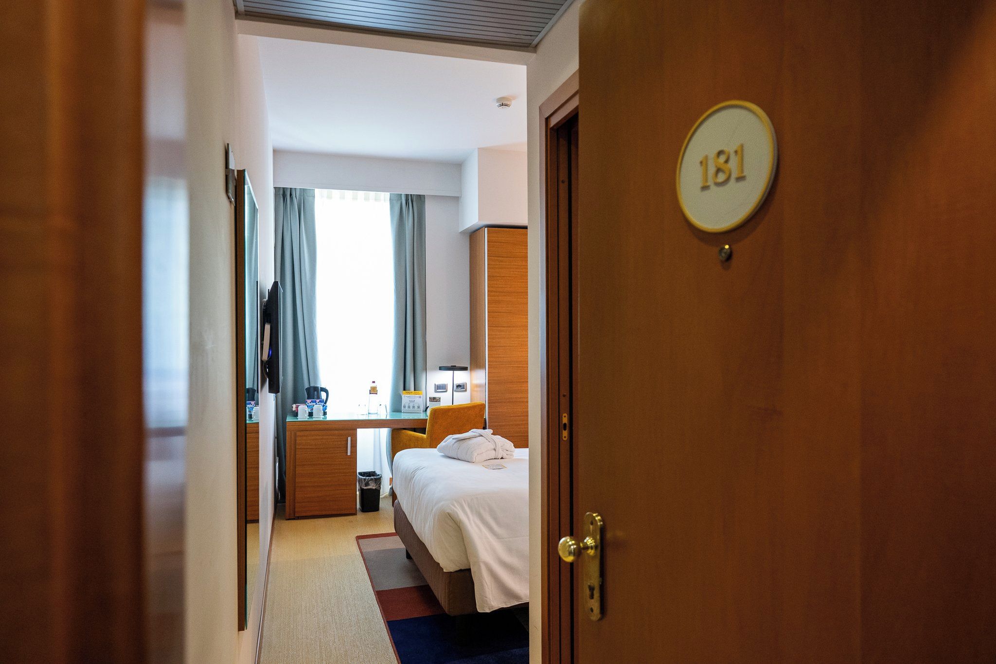 A hotel room with a bed, a desk, a chair, a mirror, and a door with a room number.