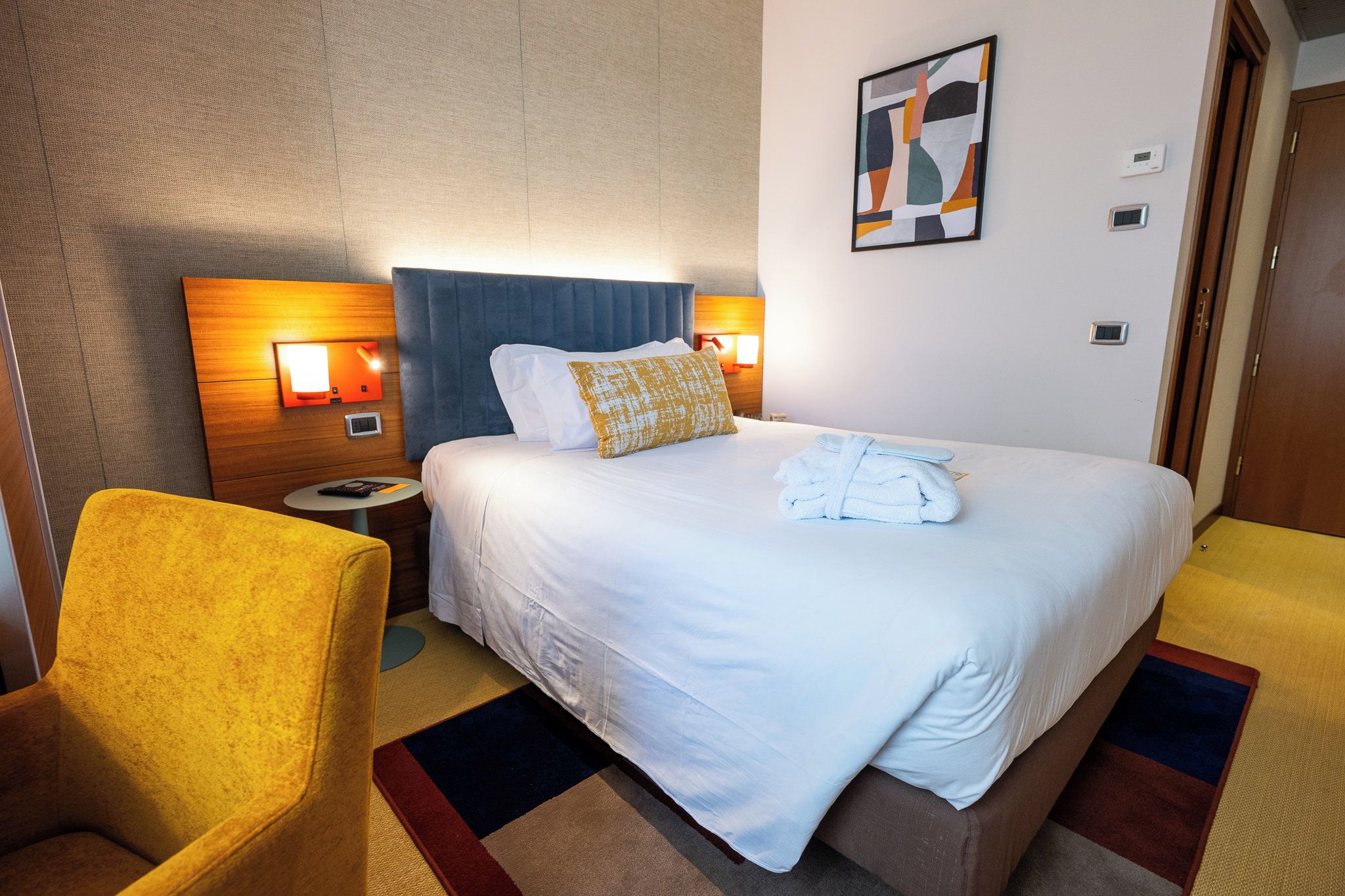 A hotel room with a bed, a nightstand, a chair, and a wall-mounted artwork.