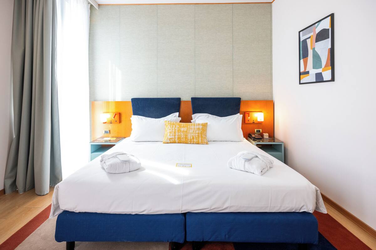 Un lit double avec une literie blanche et bleue, deux oreillers, deux tables de chevet avec des lampes, une œuvre d’art accrochée au mur et une fenêtre avec des rideaux.