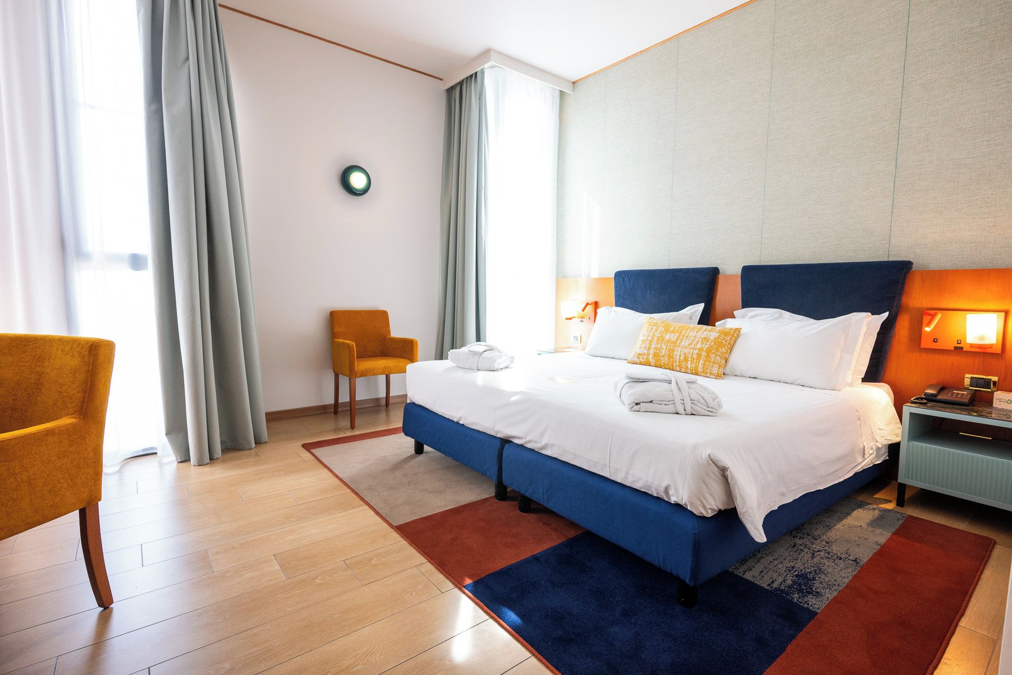 A hotel room with a bed, a chair, a bedside table, and a wall-mounted light.