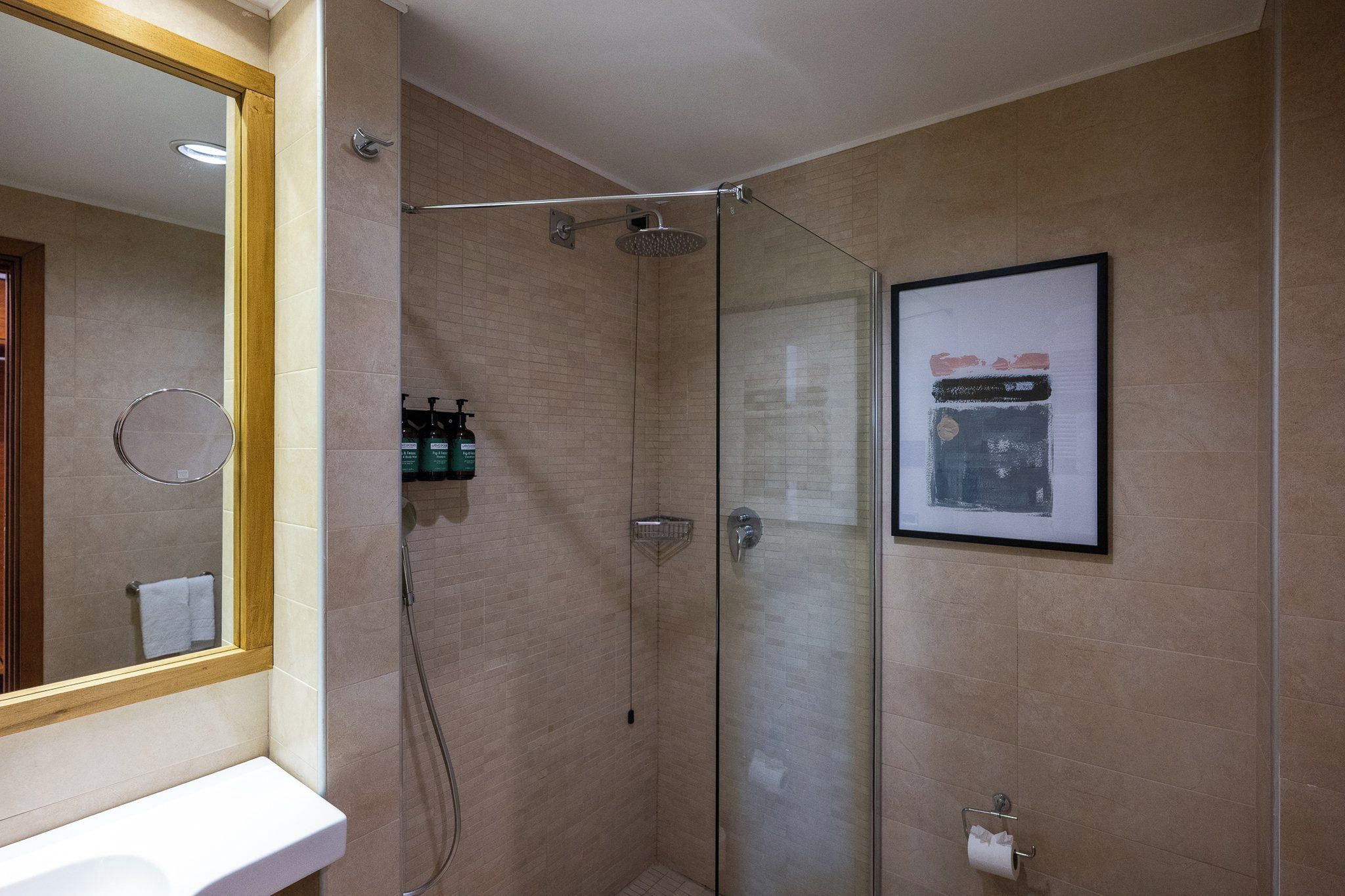 A modern bathroom with a glass shower enclosure, a framed artwork on the wall, and a mirror with a wooden frame.