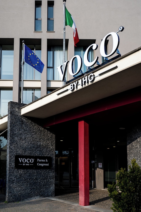 Un bâtiment sur lequel on peut lire l’inscription « VOCO BY HG », et qui est orné des drapeaux de l’Union européenne et de l’Italie.