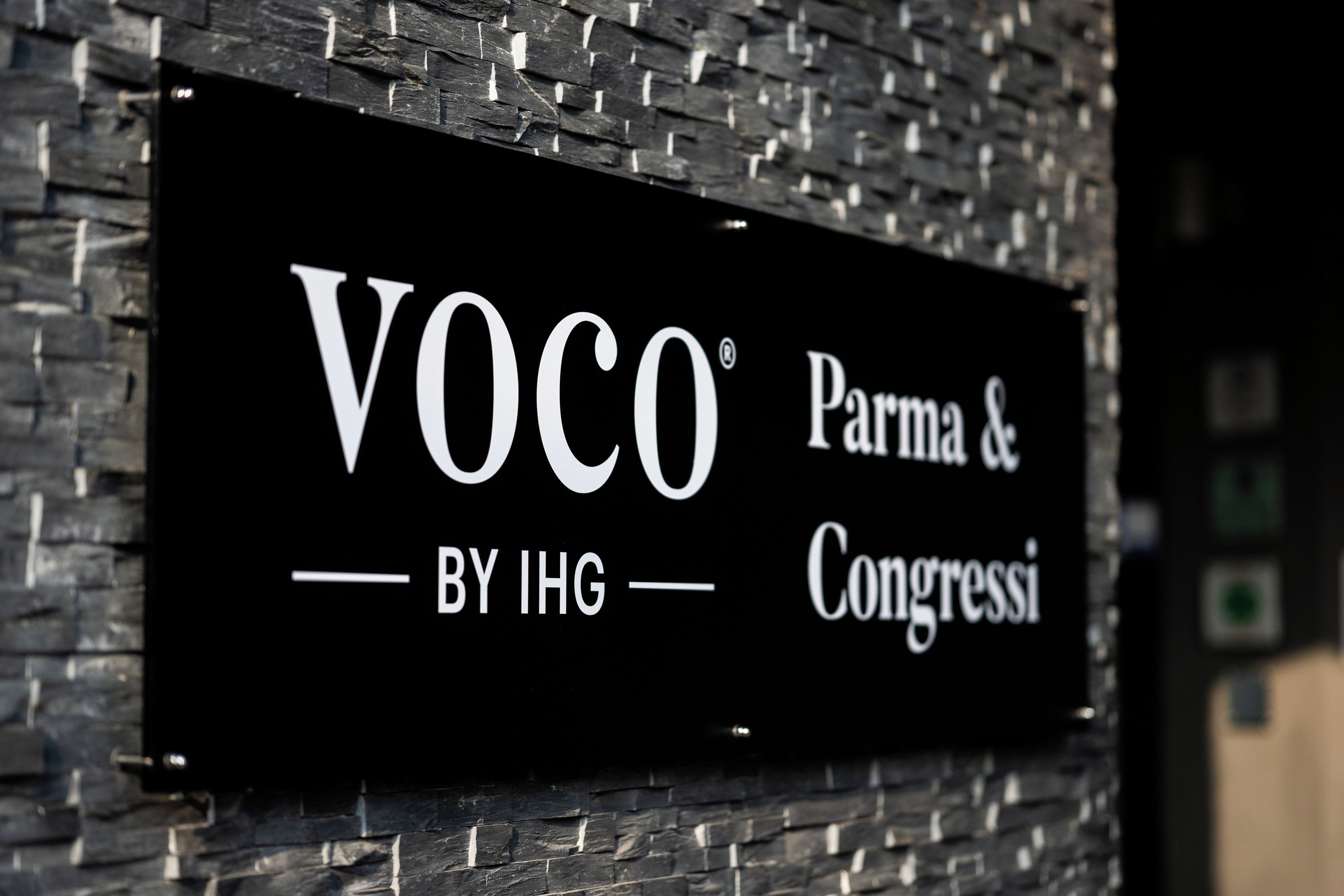 A sign with the text 'Voco by IHG' on a brick wall.