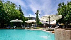 Una piscina con sillas de descanso y sombrillas, rodeada de árboles y un edificio.