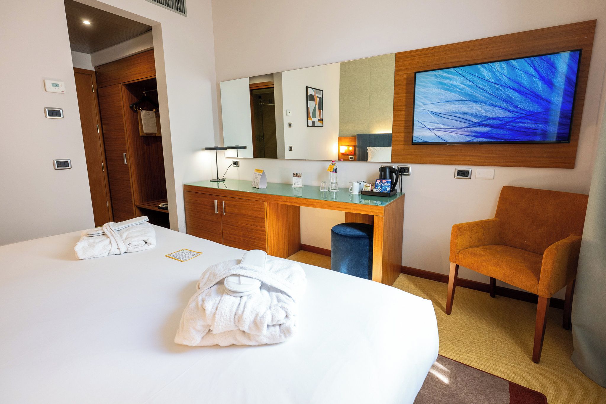 Quarto de hotel com cama, escrivaninha, cadeira, televisão e espelho.