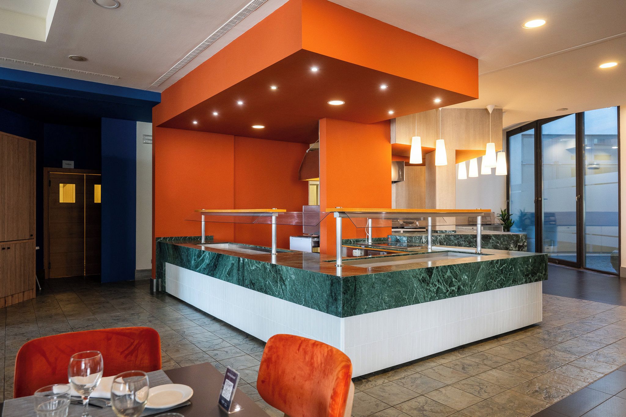 A modern restaurant with a central island, seating area, and a view through large windows.