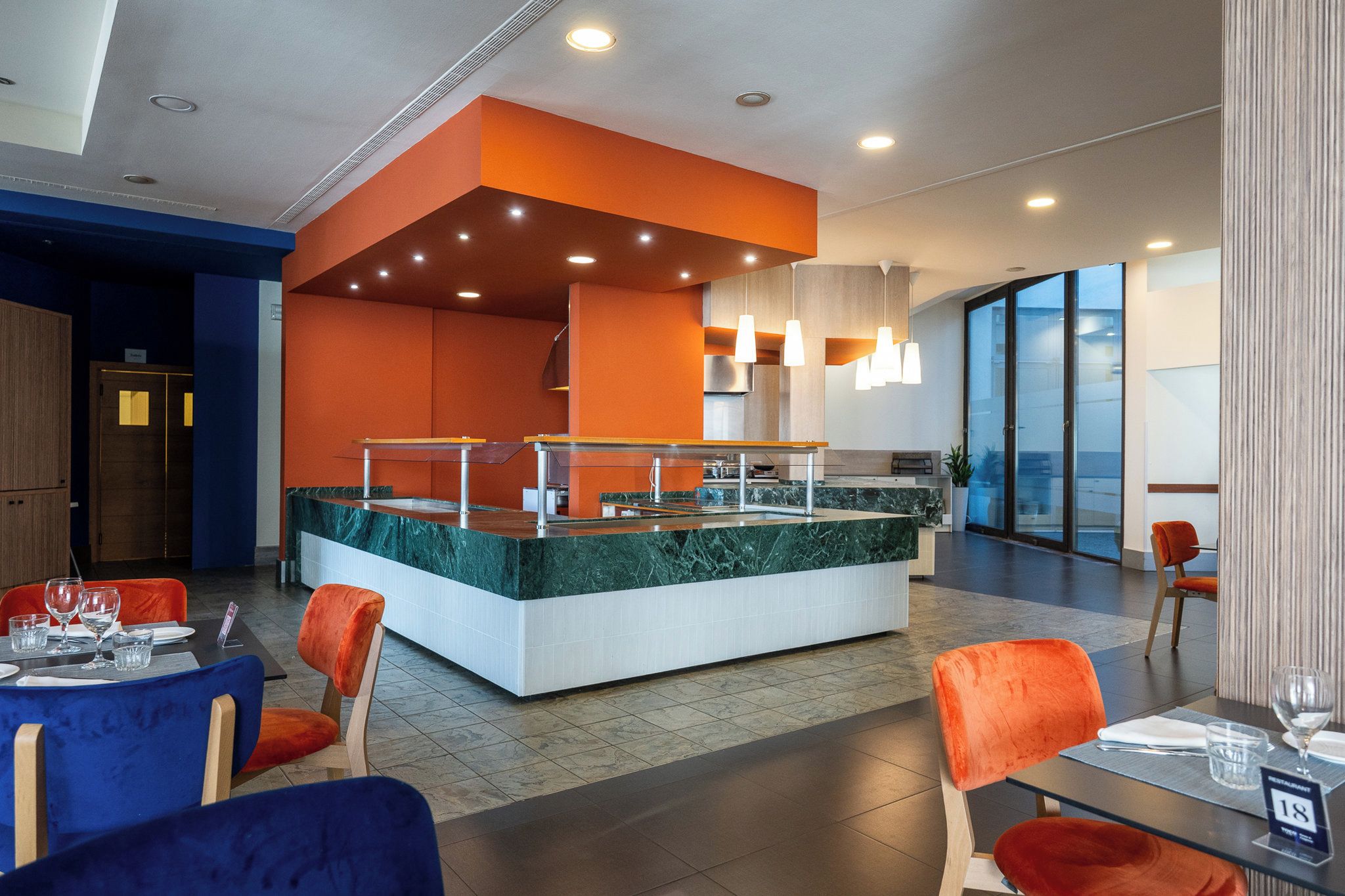 A modern restaurant with a central island counter, orange and blue chairs, and a view of the city through large windows.