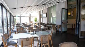 A spacious indoor dining area with multiple tables and chairs, large windows, and a modern ceiling design.