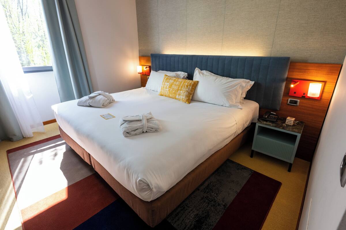Un lit bien fait, avec du linge de lit blanc et un coussin jaune, une table de chevet avec une lampe, et une vue sur l’extérieur par une fenêtre.
