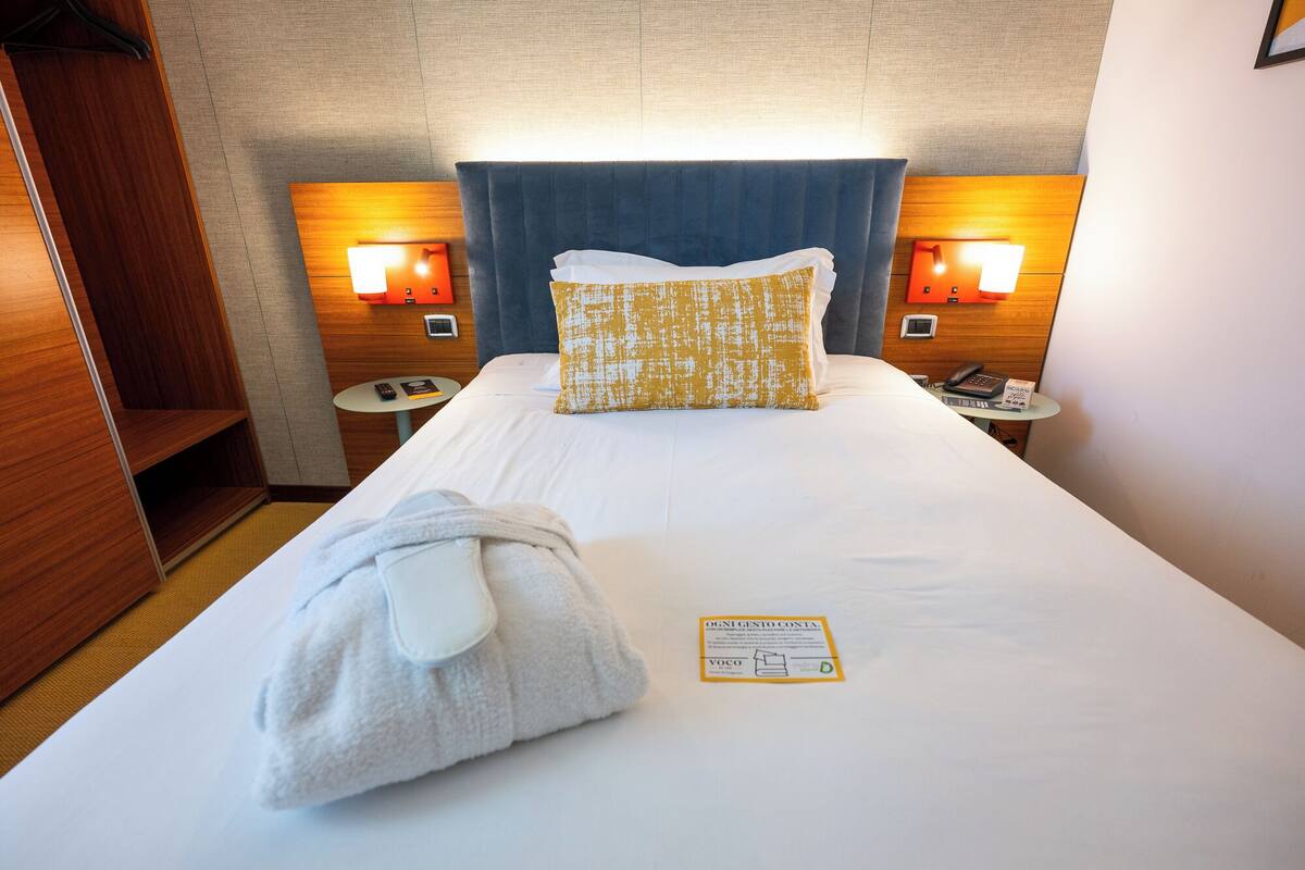 Une chambre d’hôtel comprenant un lit, un peignoir, un oreiller, une table de chevet avec un téléphone et une lampe fixée au mur.