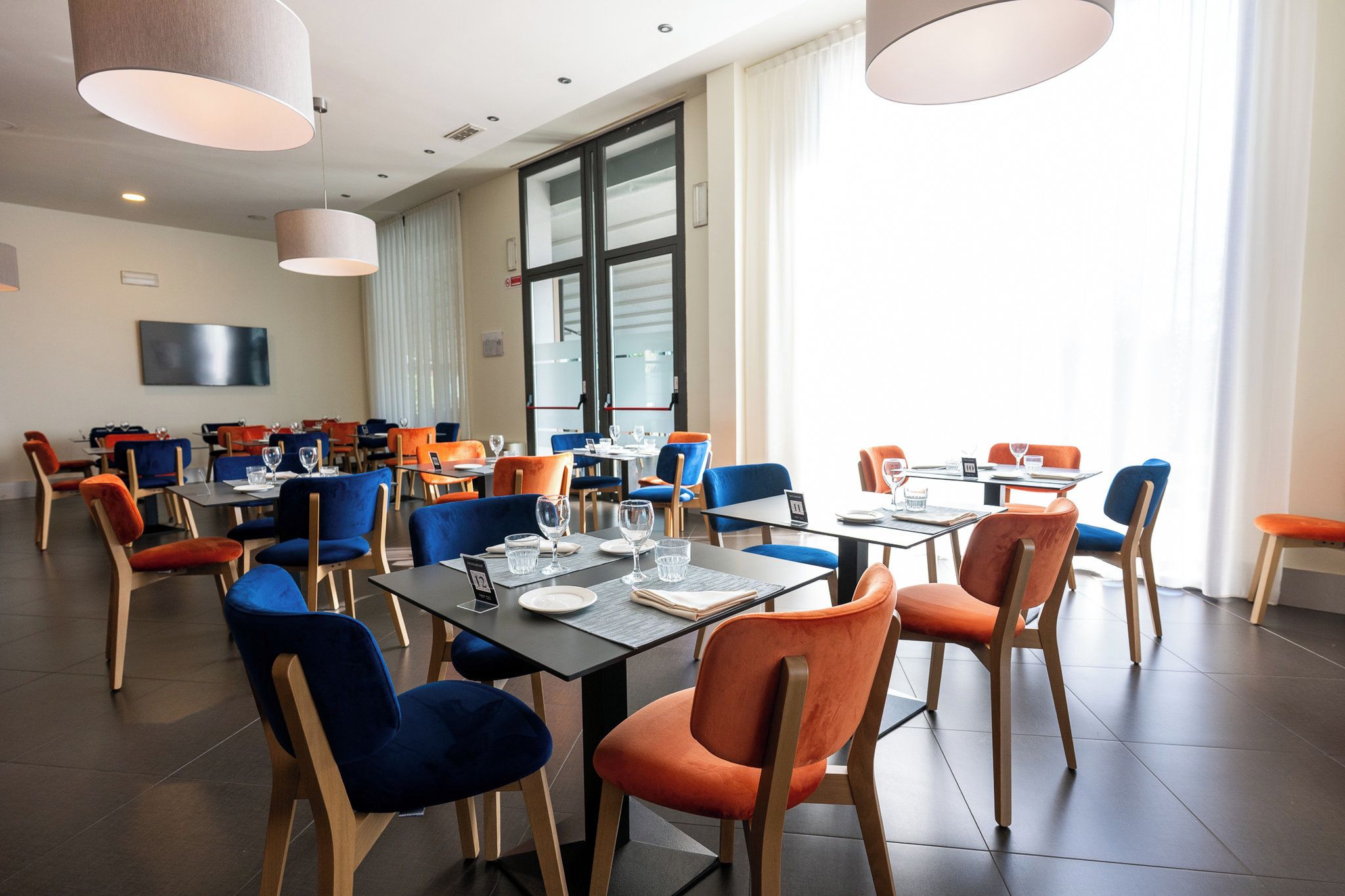 A modern dining area with tables and chairs, large windows, and a flat-screen TV on the wall.