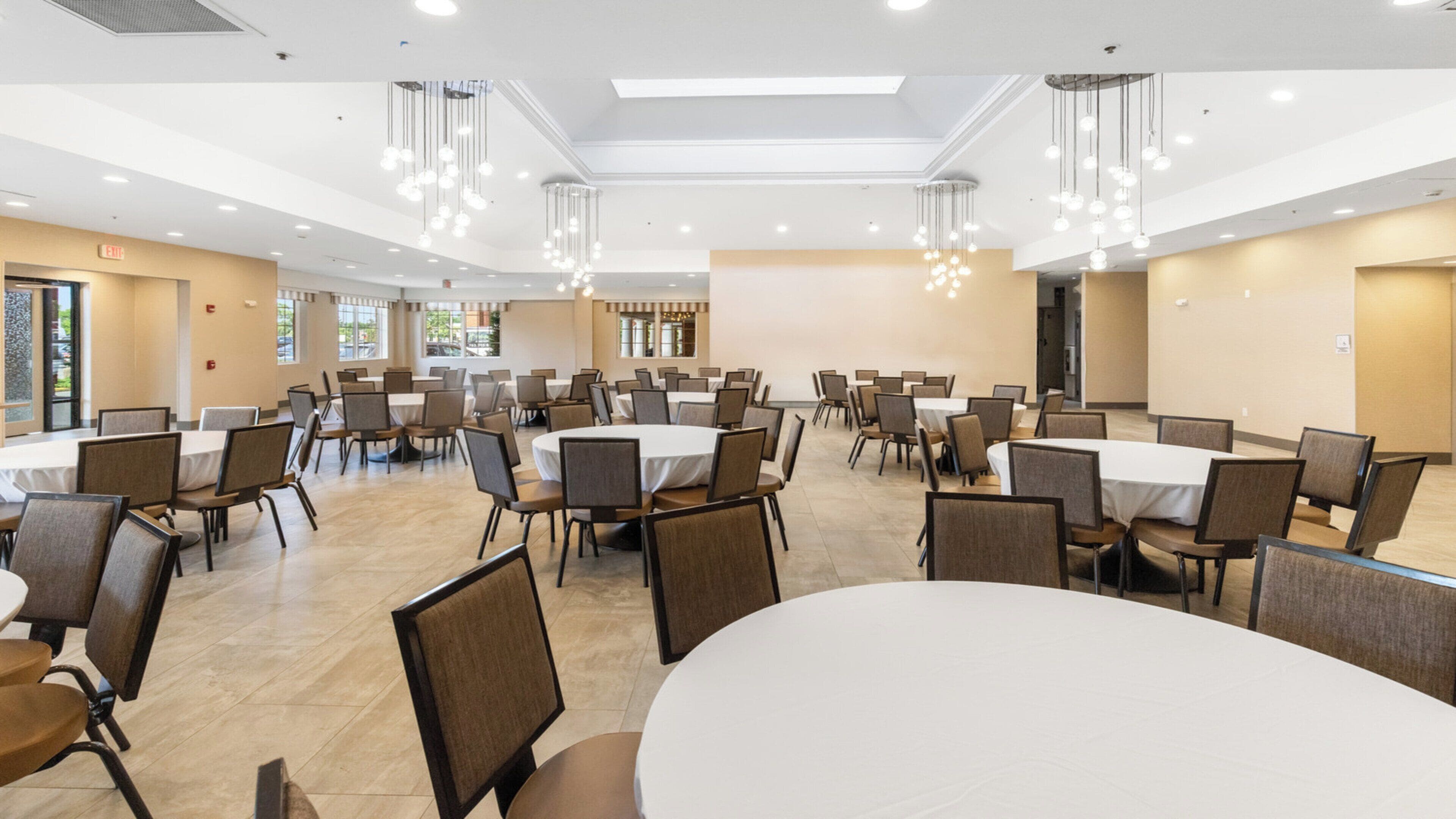 A conference room with multiple tables and chairs, large windows, and hanging light fixtures.