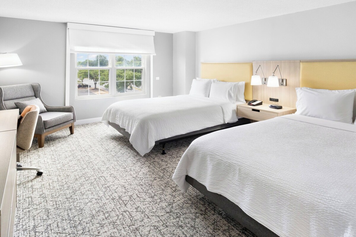 Une chambre d’hôtel avec deux lits, un bureau, une chaise, une table de chevet et une fenêtre donnant sur un paysage agréable.