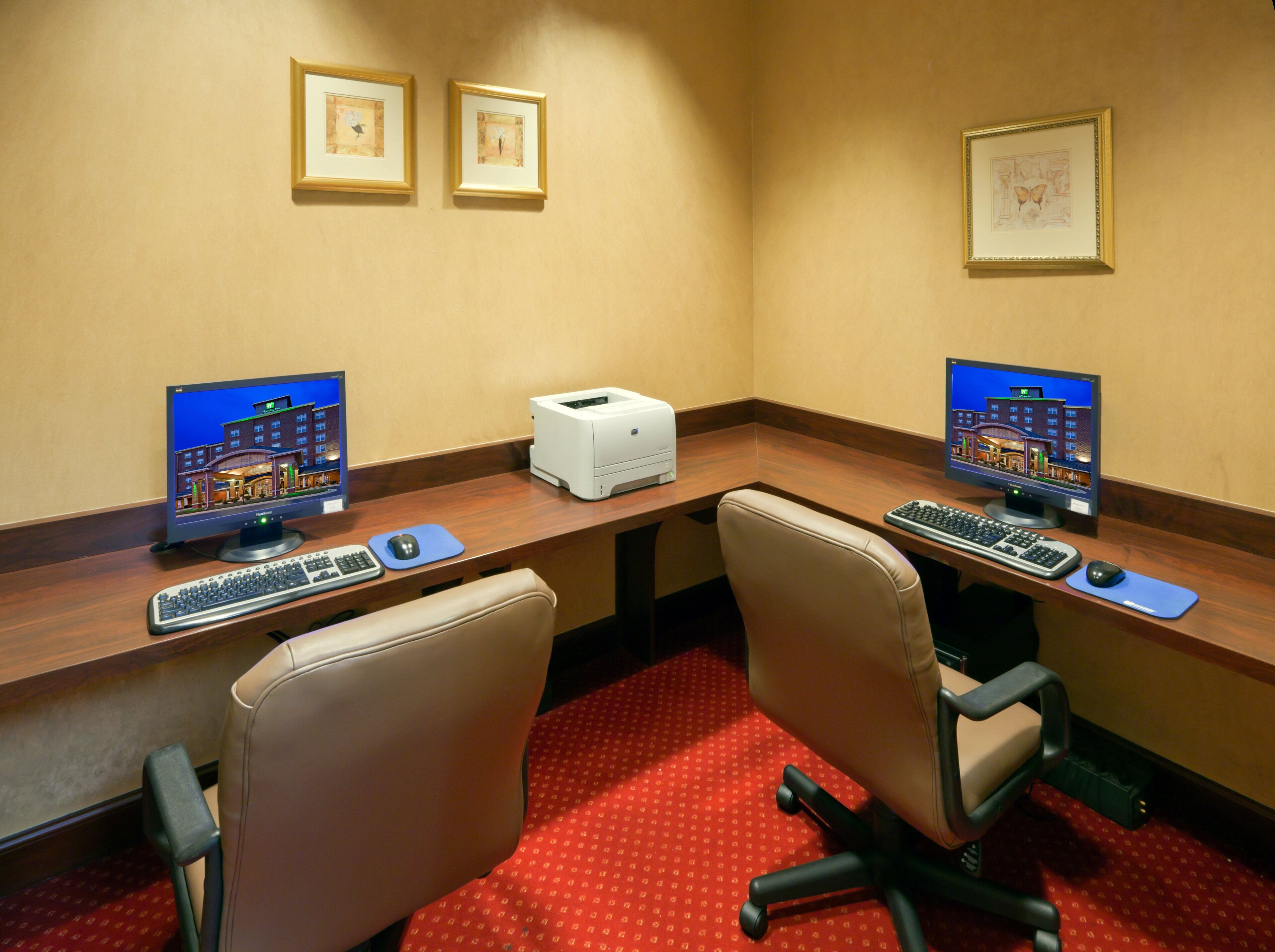 Une chambre d’hôtel avec deux bureaux, chacun équipé d’un ordinateur, d’un clavier et d’une souris. Deux tableaux encadrés sont accrochés au mur au-dessus des bureaux.