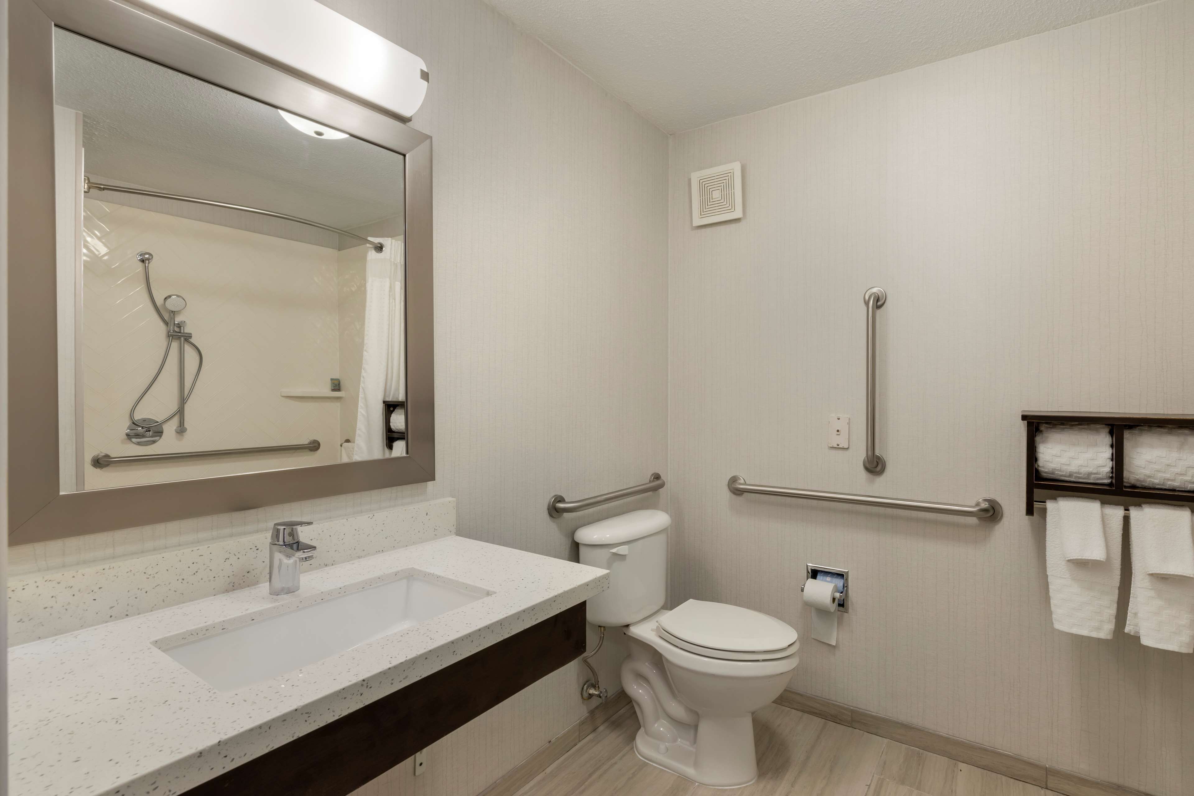 Un baño moderno con lavamanos, inodoro y ducha blancos. Incluye barras de apoyo y un perchero para toallas.