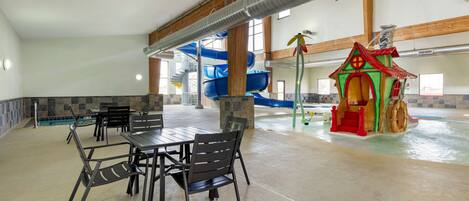 Un espace piscine intérieure comprenant un toboggan, une aire de jeux et un coin repas avec des tables et des chaises.