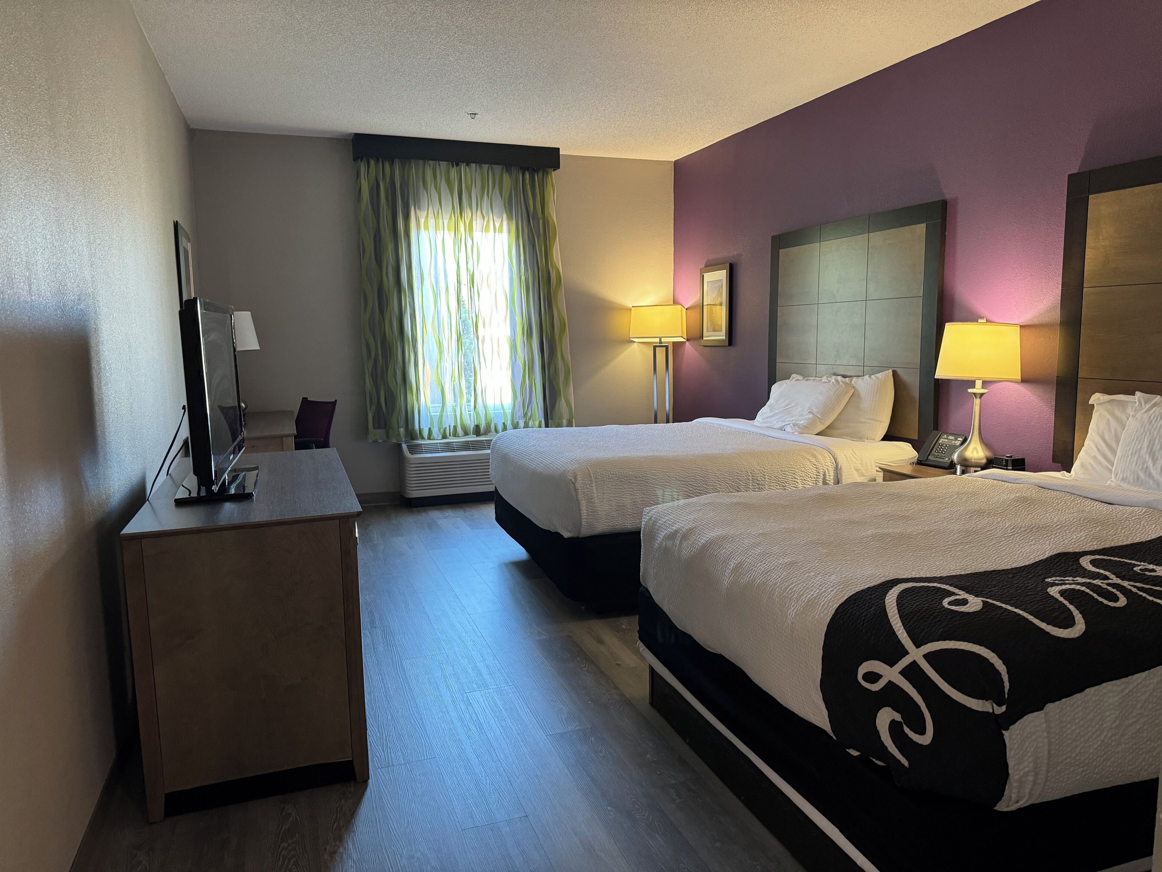 A hotel room with two beds, a desk, a flat-screen TV, a standing heater, and two bedside lamps.