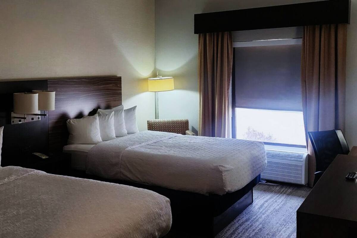 Habitación de hotel con dos camas, un escritorio, una lámpara y un ventanal grande con cortinas.