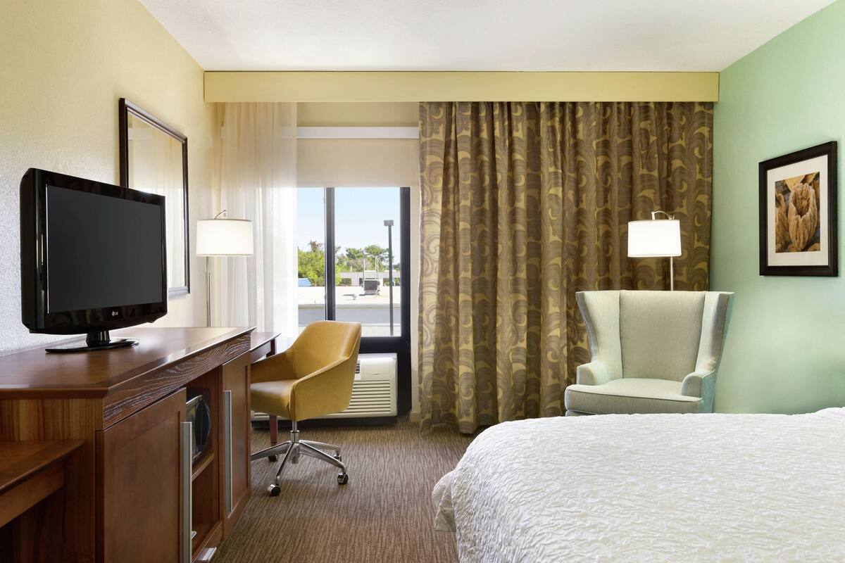 A hotel room with a bed, a desk with a TV, a chair, a lamp, and a window with curtains.