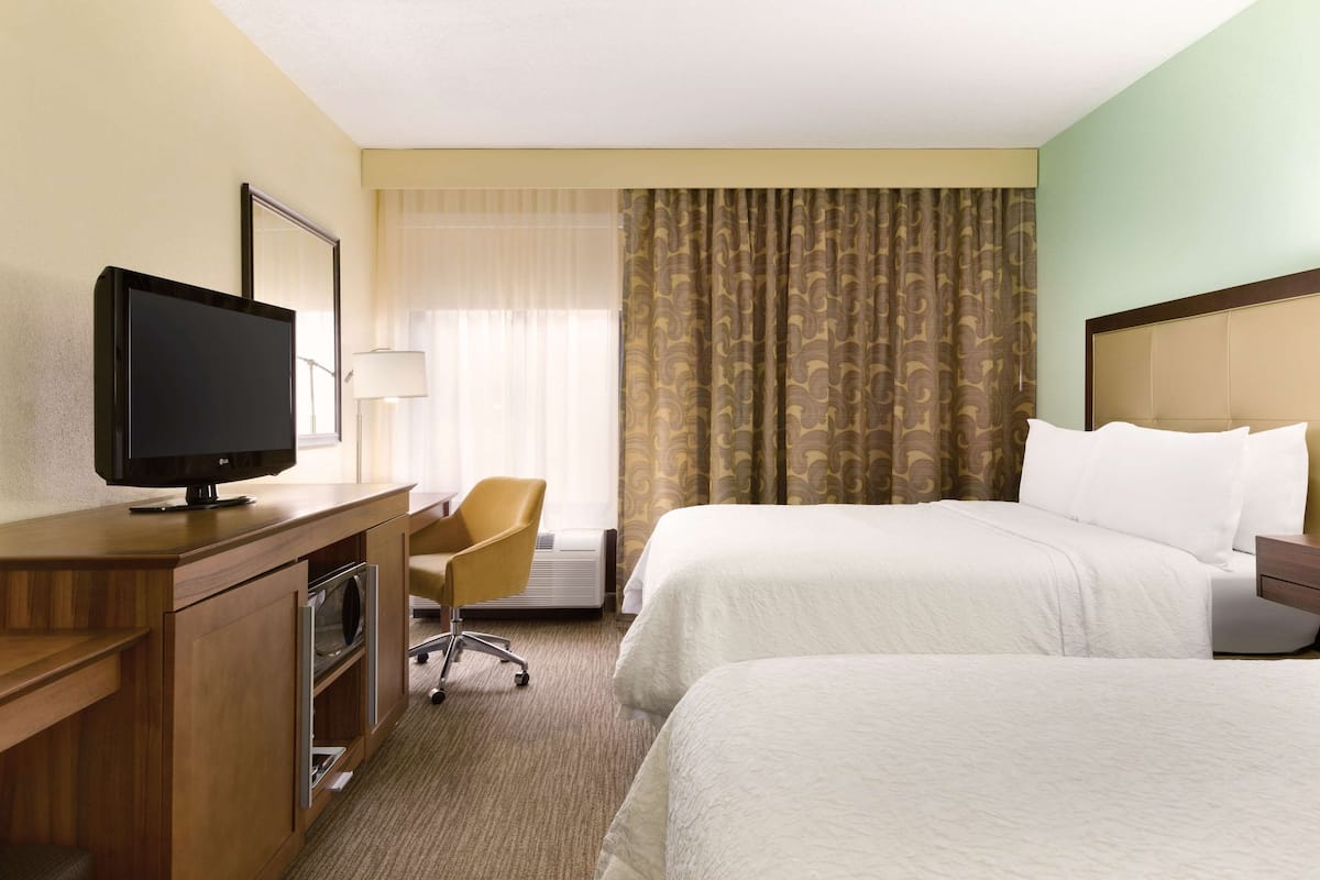 A hotel room with two beds, a desk with a television, a chair, and a window with curtains.