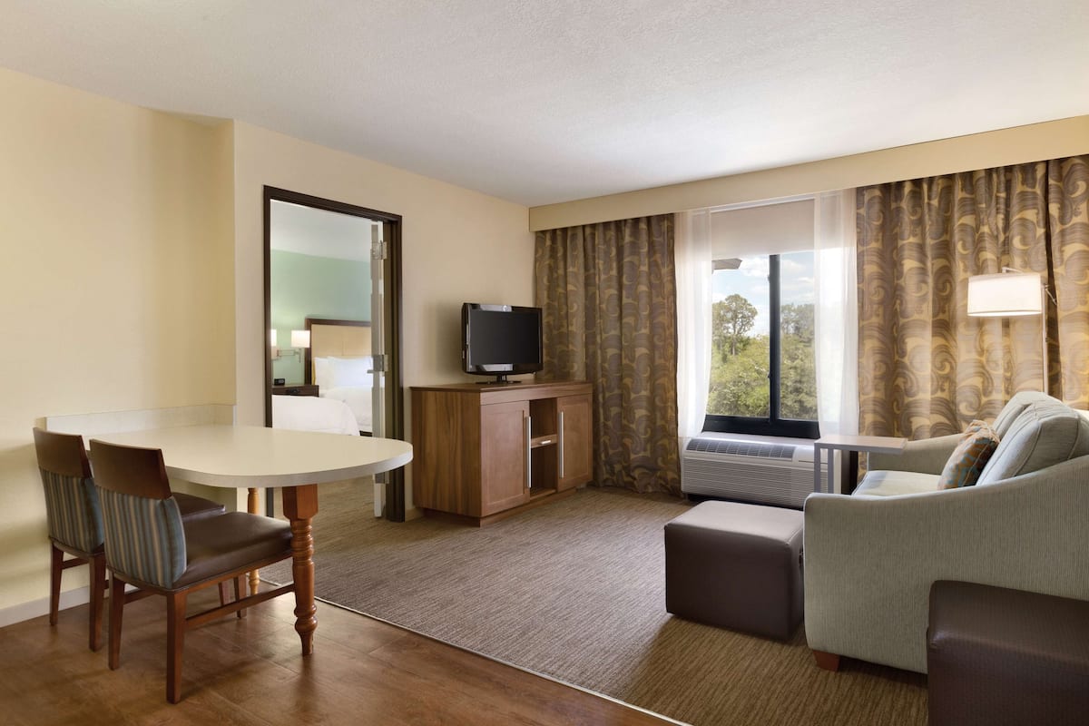 A hotel room with a dining table, two chairs, a sofa, a TV on a wooden cabinet, and a large window with curtains.