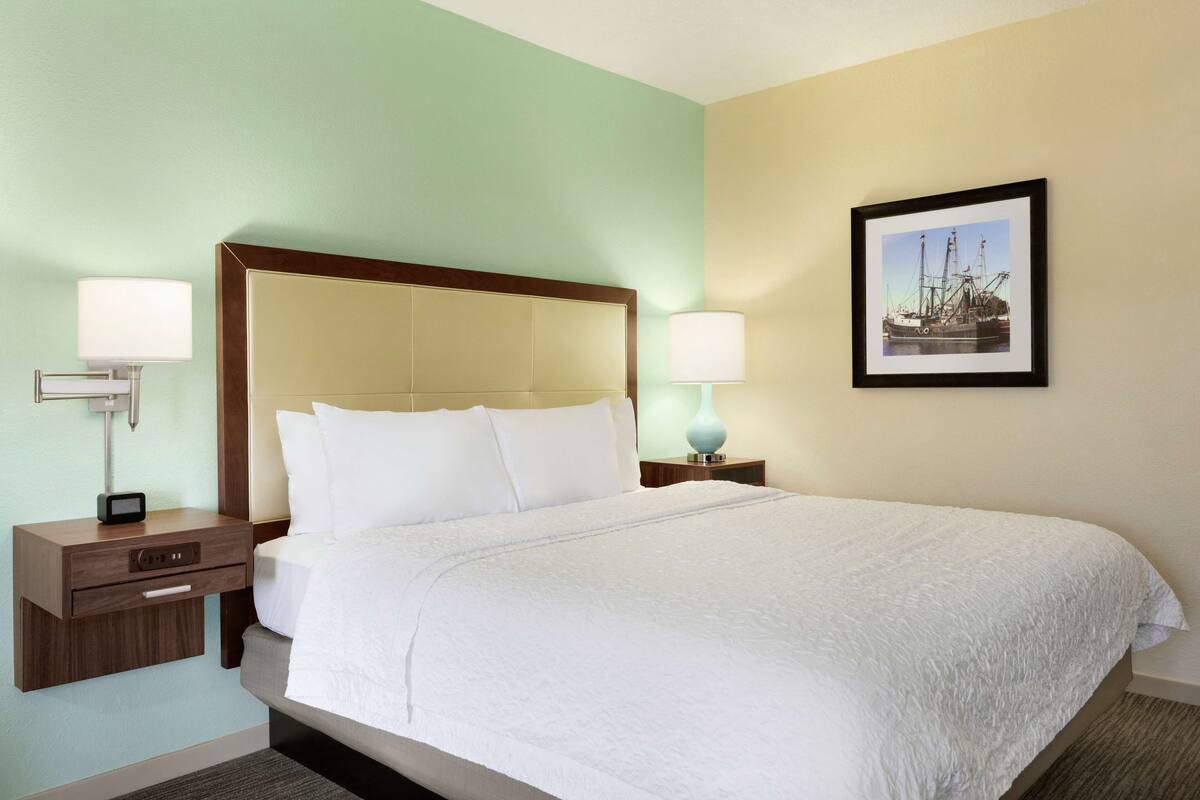 A hotel room with a large bed, a nightstand with a lamp, a framed picture on the wall, and a small table with a lamp.
