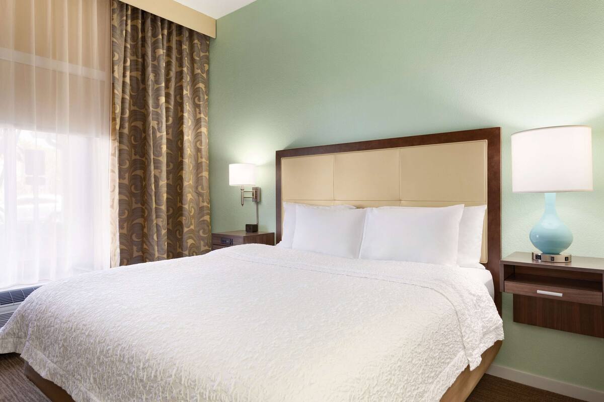 A hotel room with a large bed, a nightstand with a lamp, and a window with curtains.