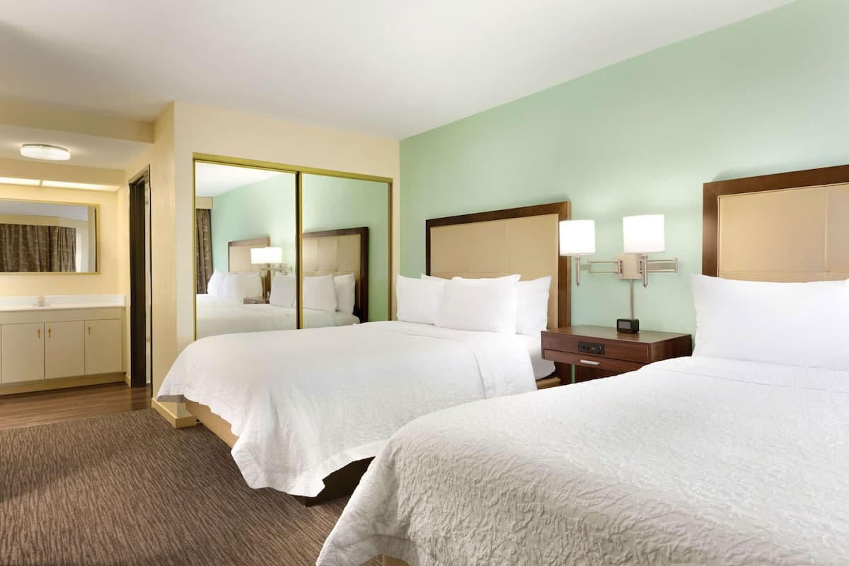A hotel room with two beds, a mirror, a nightstand, a lamp, and a bathroom with a mirror and curtains.