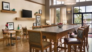 A spacious dining area with a long wooden table and matching chairs, a flat-screen TV mounted on the wall, and large windows offering a view of the outdoors. - Hampton Inn & Suites Tarpon Springs (Tarpon Springs)