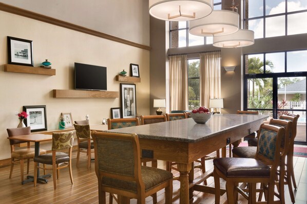 A spacious dining area with a long wooden table and matching chairs, a flat-screen TV mounted on the wall, and large windows offering a view of the outdoors. - Hampton Inn & Suites Tarpon Springs (Tarpon Springs)