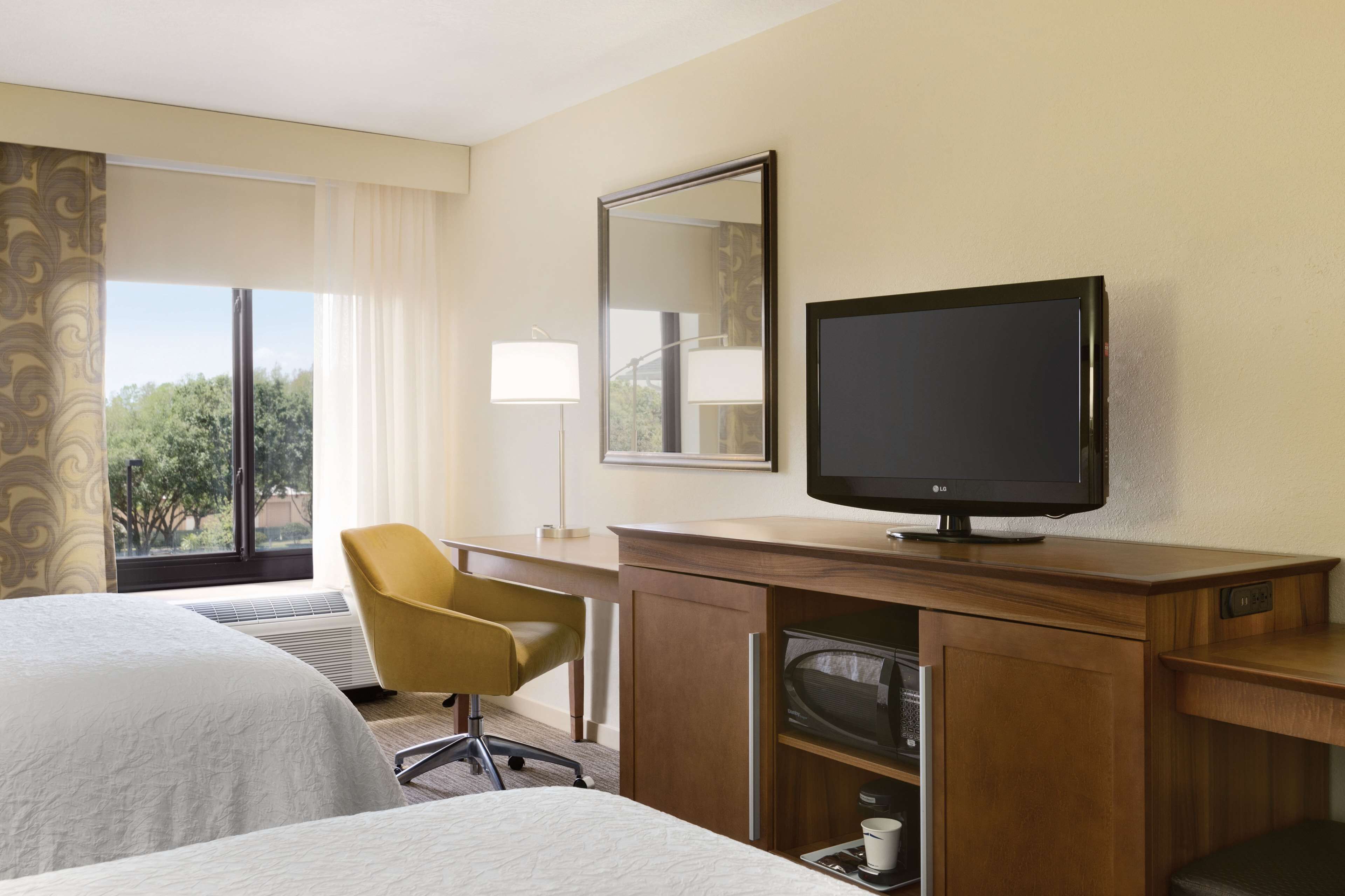 Una camera d'albergo con due letti, una scrivania con televisore, una sedia e un'ampia finestra con tende.